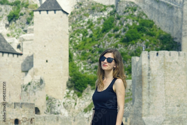 Fototapeta Portrait of attractive young woman standing in front of towers of ancient castle in Europe. Travel to most famous landmarks of Serbia: smiling female tourist enjoying walk in Golubac fortress.
