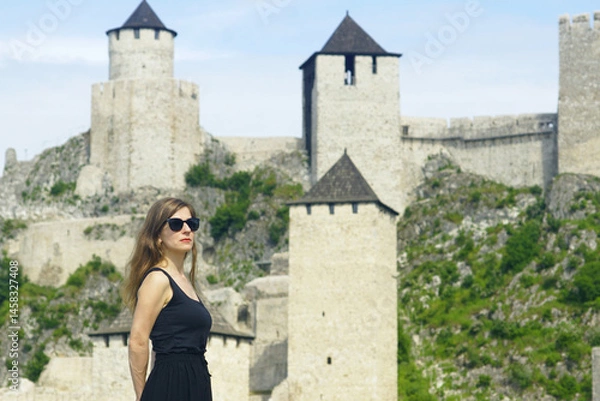 Fototapeta Woman in black clothes and sunglasses stands in front of ancient fortification. Fairytale places in Serbia: female tourist near medieval fortress Golubac with fortified towers and the keep.