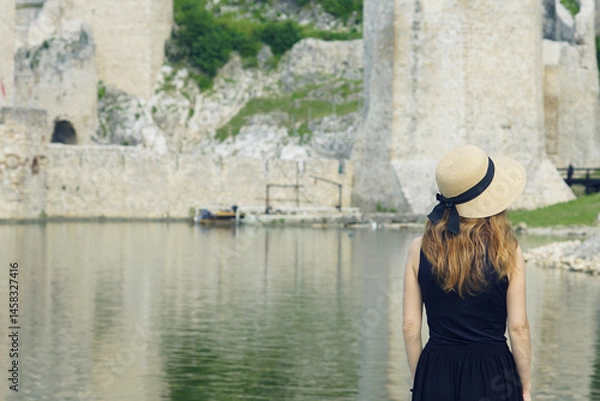 Fototapeta A woman in a hat with golden-red hair, photographed from behind, stands by the river near the walls of an ancient castle. A female tourist walks along the embankment near the Golubac fortress, Serbia