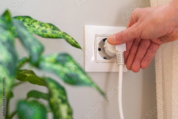 Fototapeta Close-up of a hand inserting a white electrical plug into a white wall socket next to a green houseplant. Highlighting the concepts of electricity. Power. And energy consumption in a domestic setting