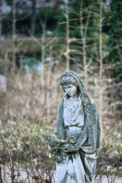 Fototapeta Moss-covered statue of a woman. Sculpture holding flowers, standing in a garden.