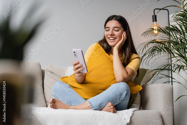 Fototapeta Digital self-care at home. Latina Indian woman in her 20s on sofa with phone, tuning into online therapy or study, blending mental wellness and digital learning in a chill home setup
