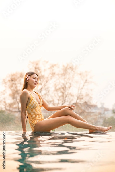 Fototapeta Asian woman in swimsuit relaxing by rooftop pool at sunset. Young tourist woman enjoying golden hour summer vibes by the pool with city skyline backdrop.