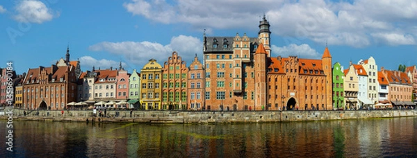 Fototapeta Panoramic view of colorful historic buildings along the Motlawa River in Gdansk, Poland. Features vibrant architecture with a backdrop of blue sky and scattered clouds