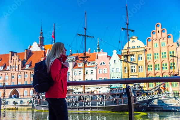 Fototapeta Tourist in red jacket with backpack walks along Gdansk waterfront. Behind her are historical buildings with vibrant facades and a traditional sailing ship at the harbor.