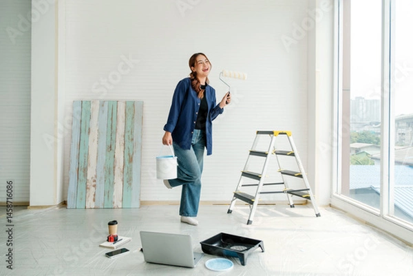 Fototapeta Smiling young woman holding a paint bucket and roller, standing in a bright room during a DIY home makeover. Surrounded by tools and laptop, expressing joy and creativity in home renovation.