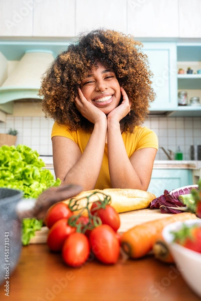 Fototapeta Happy young black woman smiling while cooking salad with fresh vegetables in the kitchen at home - Portrait picture - Food and people life style concept