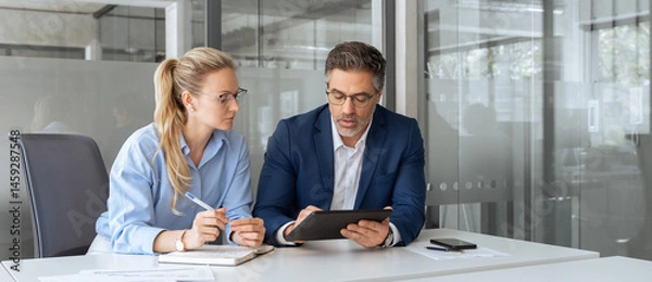 Fototapeta Team leader explain trainee working process using tablet pc computer. Two professional business people working together. Partners mature man, woman discussing project in office workplace. Copy space