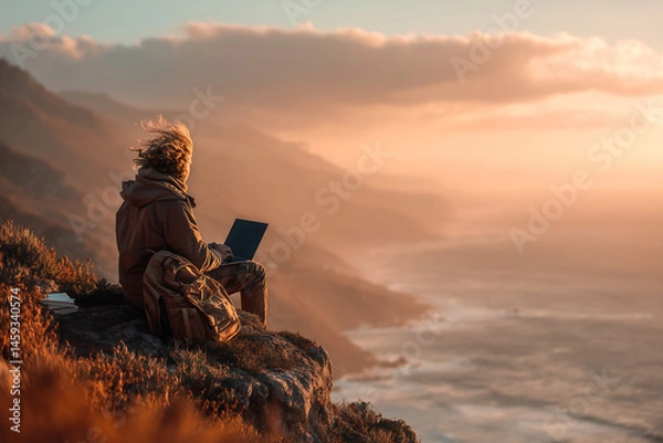 Obraz Person using a laptop on a cliffside at sunset with ocean views
