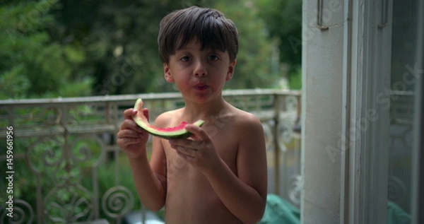Fototapeta Boy enjoying a slice of watermelon while standing on a balcony, savoring a refreshing snack in a relaxed outdoor environment, surrounded by greenery