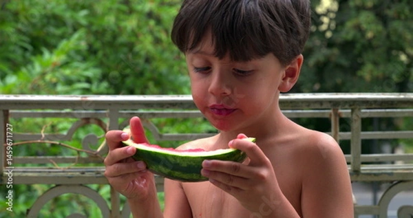 Fototapeta Young boy carefully eating a slice of watermelon, dripping juice as he enjoys the fresh and sweet fruit on a sunny day outdoors, savoring the moment