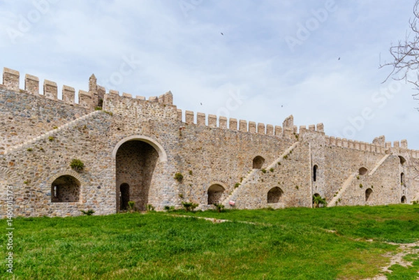 Fototapeta Historic stone castle wall featuring intricate architecture, vintage arched entryways, and a vibrant green lawn with clear skies, ideal for history and architecture enthusiasts...