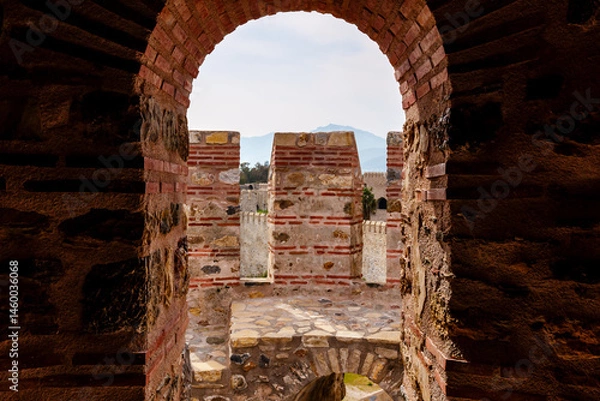 Fototapeta An antique stone fortress wall featuring a central arched gateway that frames a calming ocean view under a serene, overcast sky, symbolizing history, peace, and coastal heritage...