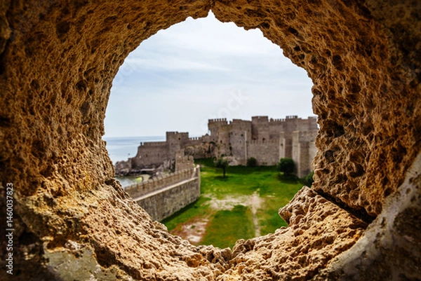 Fototapeta Magnificent Ancient Stone Castle Framed Through a Rocky Circular Opening Under Blue Sky..