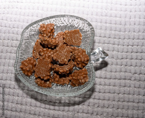 Obraz A glass dish shaped like a leaf holds an assortment of crunchy chocolate pieces. The light reflects off the surface, creating a tempting display on a soft, textured background