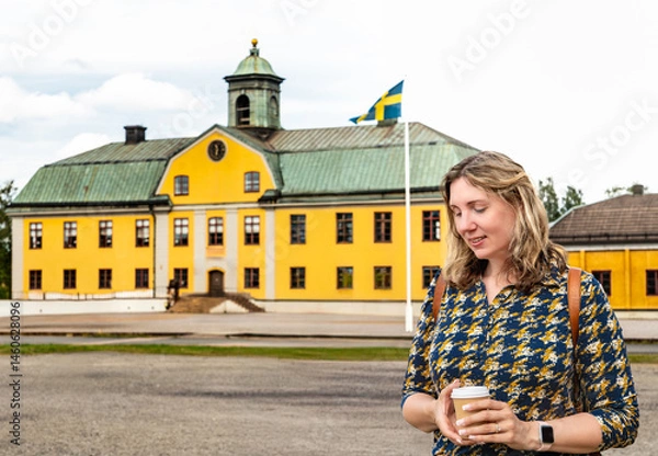 Fototapeta A woman with dark blonde hair smiles down at her coffee cup while standing in front of a charming old yellow building in Sweden, adorned with the Swedish flag
