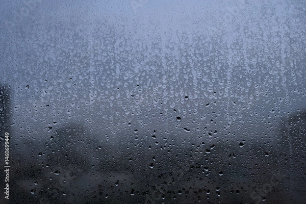 Fototapeta Raindrops on a glass window with a blurry cityscape in the background on a cloudy day.