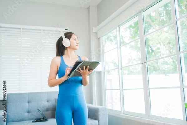 Fototapeta A focused woman in blue activewear intently uses a tablet, possibly for a workout routine, in a bright, modern home setting with a soft grey couch and natural light filtering through nearby blinds