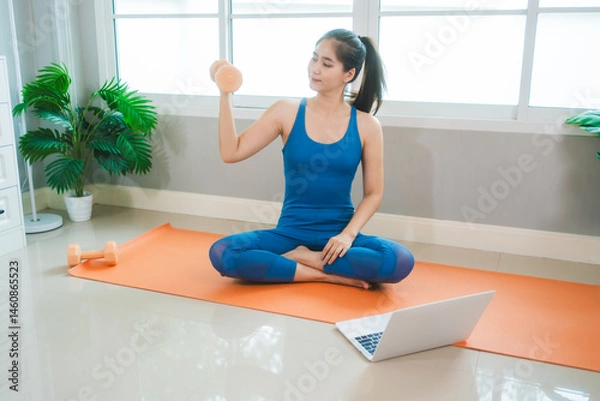 Obraz Young Asian woman practices yoga by stretching her back on a yoga mat in the living room at home. She performs pilates with challenge and motivation for health, fitness, and wellbeing