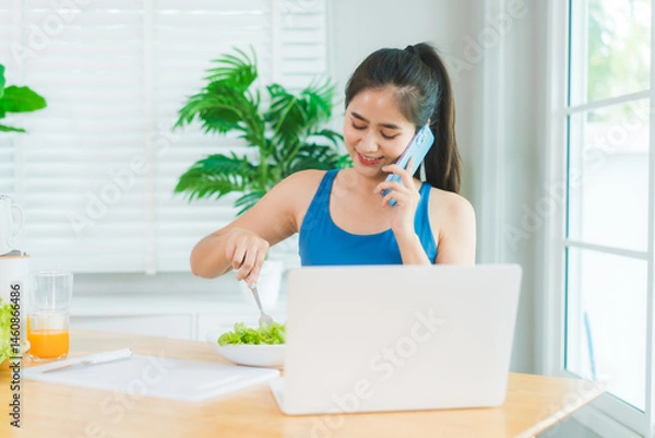 Obraz Beautiful Asian woman in fitness sportswear happily smiles while slicing vegetables and fruits on cutting board.She focuses on health nutrition at home,offering online guidance through her computer
