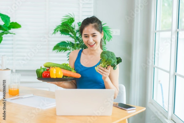 Obraz Beautiful Asian woman in fitness sportswear happily smiles while slicing vegetables and fruits on cutting board.She focuses on health nutrition at home,offering online guidance through her computer