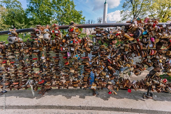 Obraz Close-up of a bridge railing densely covered with love padlocks in Riga, symbolizing eternal love, with the Freedom Monument faintly visible behind..