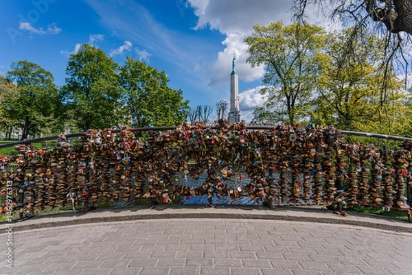 Obraz Bridge railing in Riga covered with love padlocks, symbolizing romance and commitment, with the Freedom Monument in the background on a sunny day..