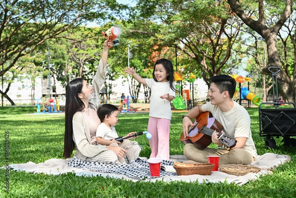 Obraz A family of four is enjoying a picnic in a park. The man is playing guitar while the children are eating. Scene is happy and relaxed.