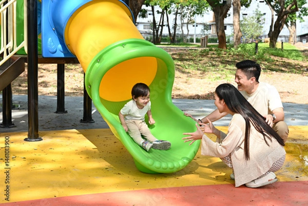 Obraz A family is standing in front of a slide. The child is sitting on the slide while the parents look on. Scene is happy and playful.