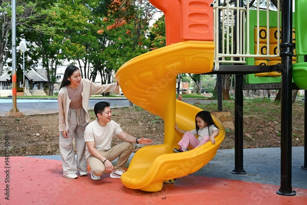 Fototapeta A family is standing in front of a slide. The child is sitting on the slide while the parents look on. Scene is happy and playful.