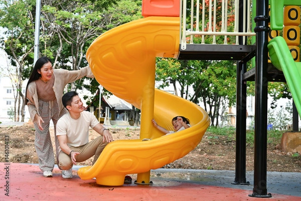Obraz A family is standing in front of a slide. The child is sitting on the slide while the parents look on. Scene is happy and playful.