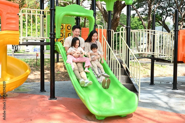 Fototapeta A family of four is enjoying a day at the park, riding a green slide together. Scene is happy and playful, as the family is spending quality time together and having fun.