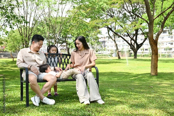 Obraz A family of four sits on a park bench, enjoying a sunny day together. The children are playing and the parents are watching over them.