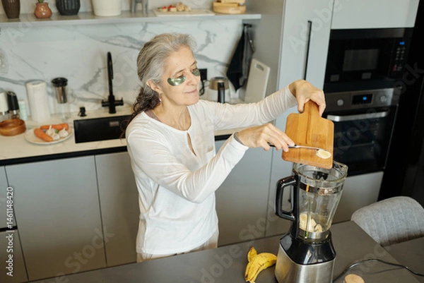 Fototapeta Senior woman with eye patches making a smoothie in a modern kitchen with various kitchen gadgets. She is wearing white clothes and appears focused on adding ingredients to blender
