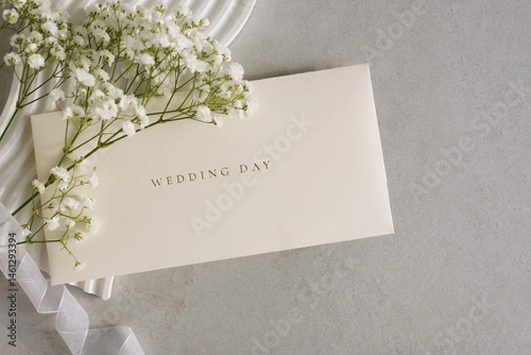 Fototapeta Wedding greeting card with a white flowers , ribbon, decor on light beige textured background. Wedding invitation with envelope.Copy space.