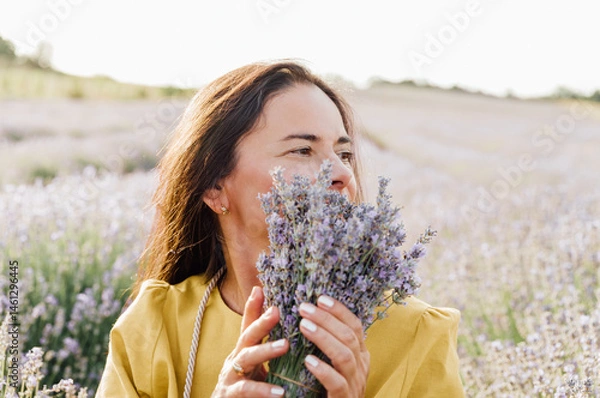 Fototapeta woman in yellow dress stands in lavender field, gently holding bouquet of lavender flowers. She appears serene and contemplative, enjoying beauty and fragrance of blossoms around her.