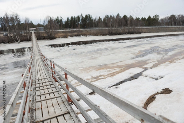 Obraz A wooden bridge spans a tranquil water body, surrounded by snow