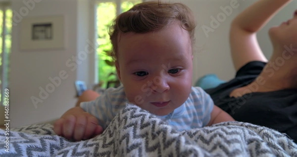 Fototapeta Baby lying on a blanket, looking slightly frustrated and emotional, in a warm home setting with natural light, while mother observes in the background