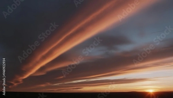 Fototapeta Dramatic clouds streak across a dusky sky. The setting sun casts a warm glow, contrasting with the dark expanse of the landscape below.