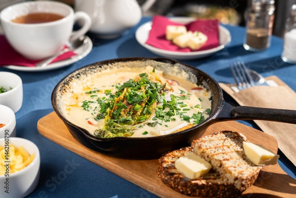 Fototapeta Baked scrambled eggs with goat cheese and vegetables served in a cast iron skillet with dark whole wheat bread, chives, yellow cheese and greens. A breakfast set, illuminated by the morning sun.