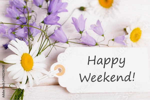 Fototapeta Happy weekend message on white gift tag with daisies and violet bellflowers on rustic wooden background, floral greeting card design, top view composition for summer celebration or weekend wishes