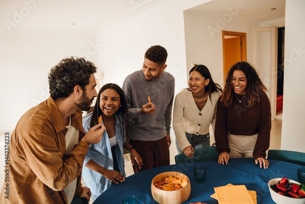 Fototapeta A group of young multiracial friends standing around the table eating snacks and laughing indoors at home.