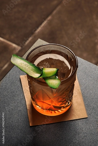 Fototapeta Crystal cocktail glass with cucumber garnish on bar top