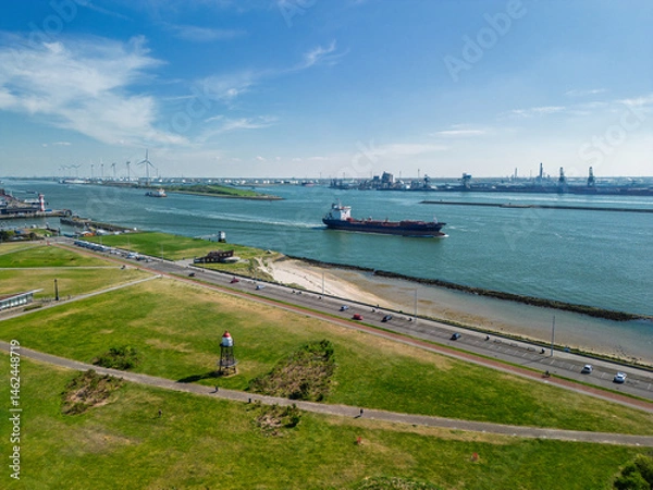 Fototapeta Aerial view of cargo ships navigating a wide river or channel near an industrial port with cranes and smokestacks in the background under a clear blue sky.