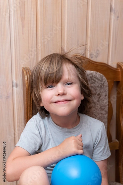 Fototapeta Blue-eyed boy blowing up a blue balloon while sitting on a wooden chair on a wooden background