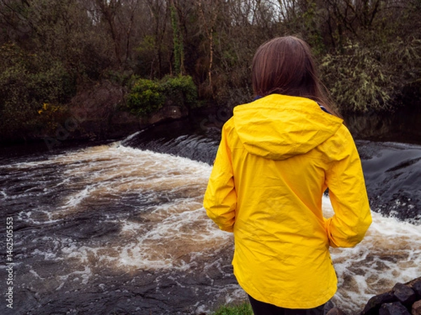 Fototapeta Female tourist in yellow jacket looking at a small river with waterfall. Travel and tourism. Enjoy nature scenery theme. Girl standing by a river with nice view.