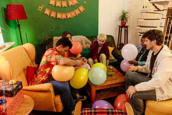 Fototapeta Group of five friends sitting at a table and blowing up balloons, at a party