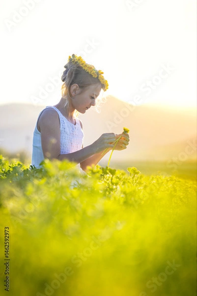 Fototapeta Young joyful girl on a blooming meadow with a wreath of dandelions on her head at sunset. Spring joyful mood child with flowers in hand