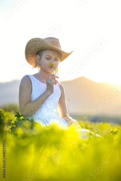 Fototapeta Young joyful girl on a blooming meadow with a hat on her head at sunset. Spring joyful mood child in the grass with dandelion in the hand