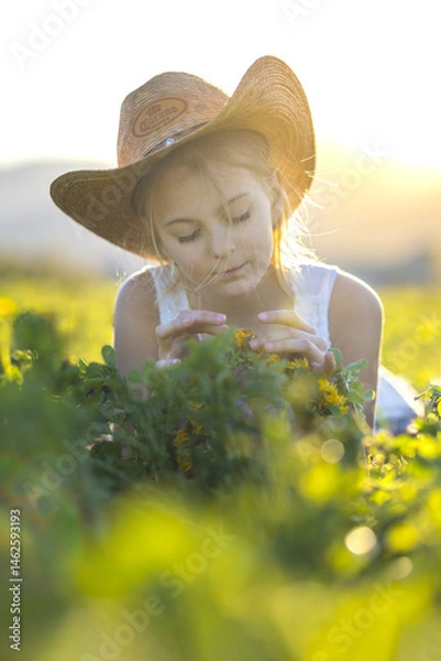 Fototapeta Young joyful girl on a blooming meadow with a hat on her head at sunset. Spring joyful mood child in the grass